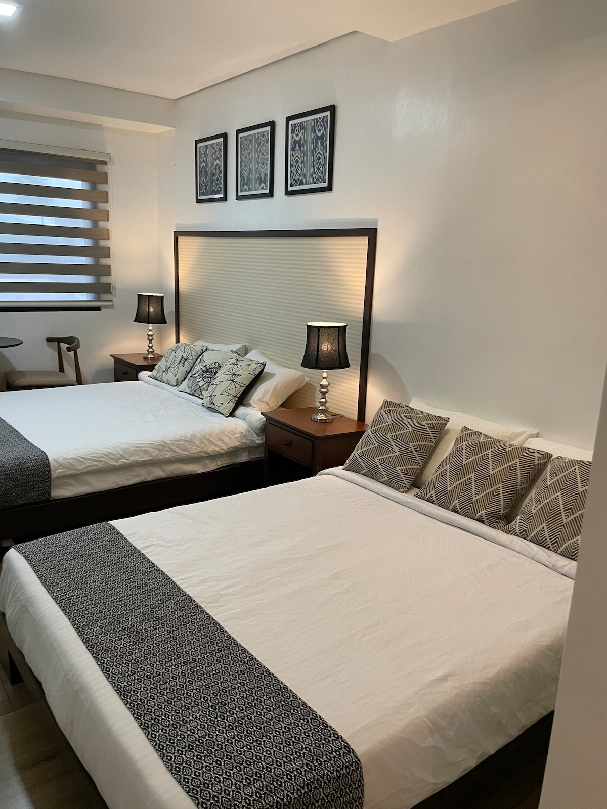 A well-appointed room featuring two spacious beds with patterned bed linens and decorative pillows. Nightstands with lamps are positioned beside each bed. Wall art and a window with layered blinds provide an elegant touch to the serene environment.