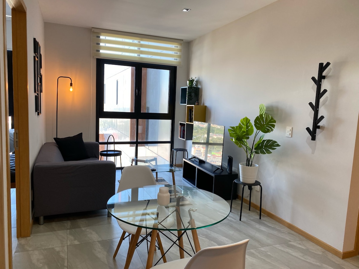 A modern living space is shown, featuring a gray sofa next to a large window. A round glass table with wooden legs is positioned in the foreground. Green foliage is displayed in a white planter, and a black shelving unit is visible against the wall.