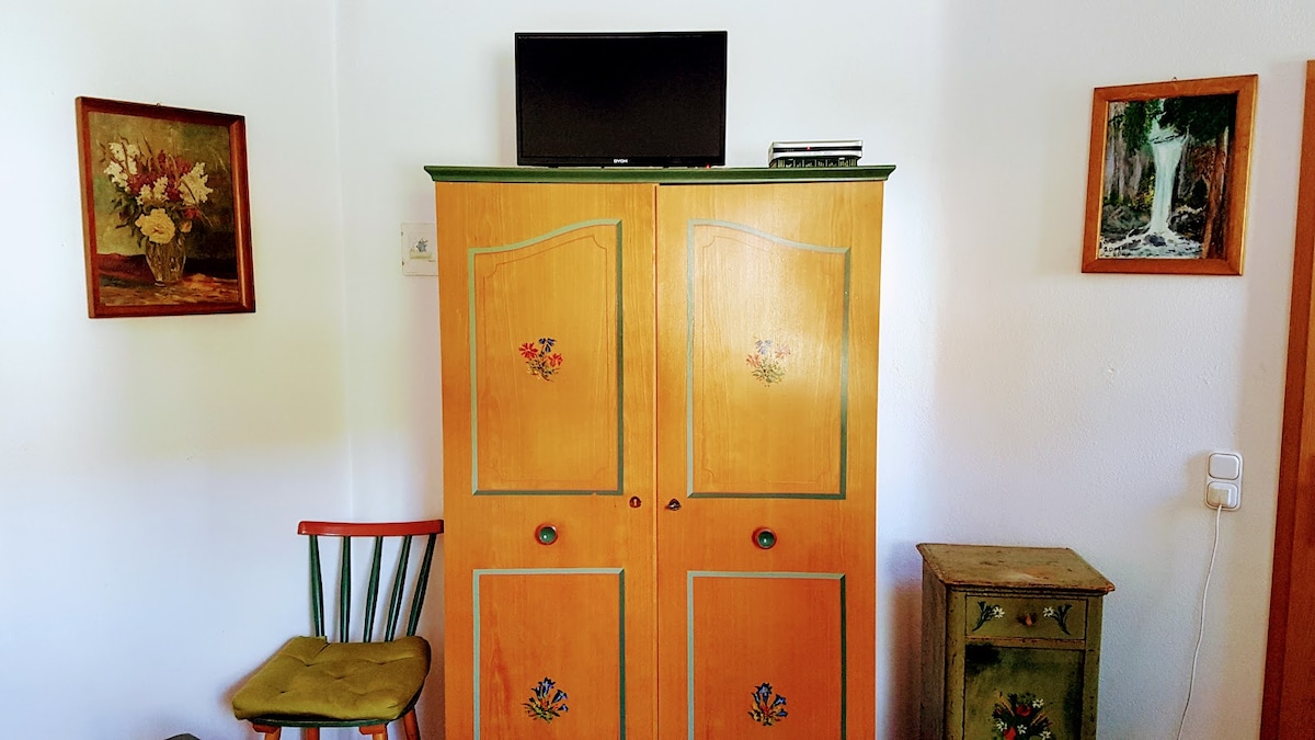 A wooden cabinet with painted floral designs stands prominently, housing a television on top. Two framed artworks with nature-themed images adorn the walls, while a green chair with a yellow cushion and a small side table are also present in the space.