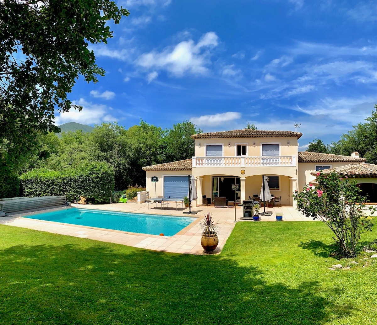 A spacious villa is set against a backdrop of lush greenery, featuring a large swimming pool framed by a neatly maintained yard. Sun loungers and a patio table are visible near the pool, while the two-story structure showcases an outdoor patio area and manicured landscaping.