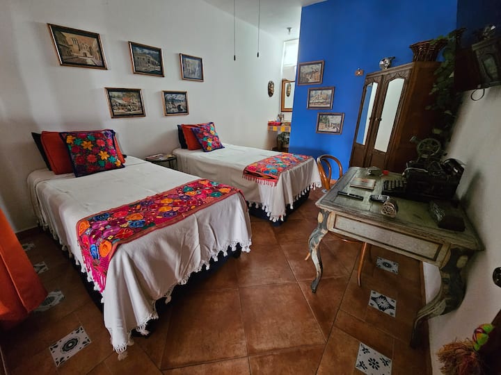 Recámara con dos camas individuales,  ropero antiguo, pantalla, minisplit, baño completo y 1 escritorio y objetos antiguos y esta recamara  tiene cuadros representativos de Monterrey de finales del siglo XIX.