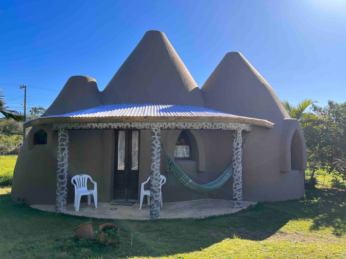 The exterior of a unique dome-shaped house is displayed, featuring three conical roofs. A small porch is shown, furnished with a white chair and a hanging hammock. Natural stone columns support the roof, and greenery surrounds the structure, creating a serene setting.