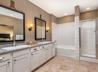 A spacious bathroom features dual vanities with large mirrors and modern sconces. A glass shower enclosure stands next to a relaxing bathtub. The neutral tile flooring complements the light cabinetry and granite countertops.