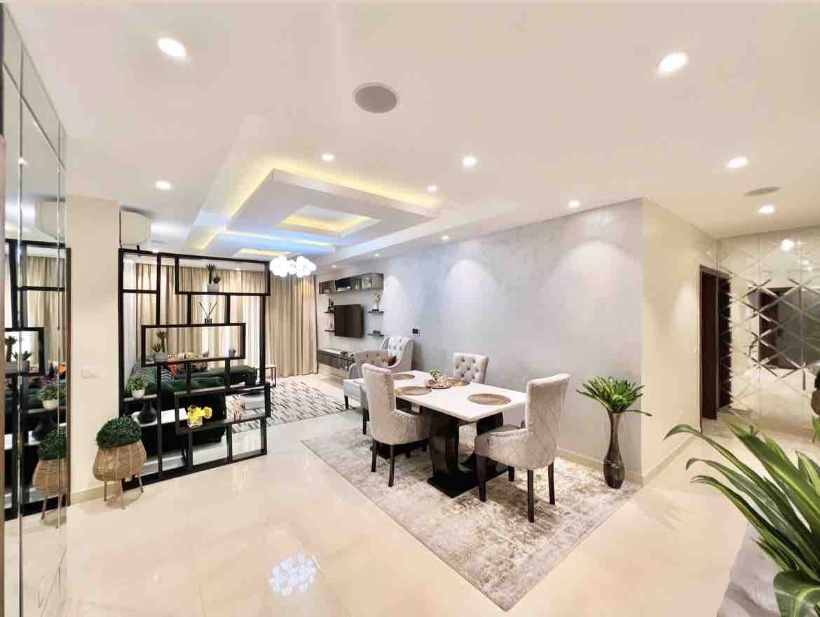 A modern living space features a dining area with a round table surrounded by four upholstered chairs. A patterned rug lies beneath, and decor elements include shelves displaying plants. Bright overhead lighting and a neutral palette enhance the airy atmosphere, complemented by a sleek wall-mounted TV.