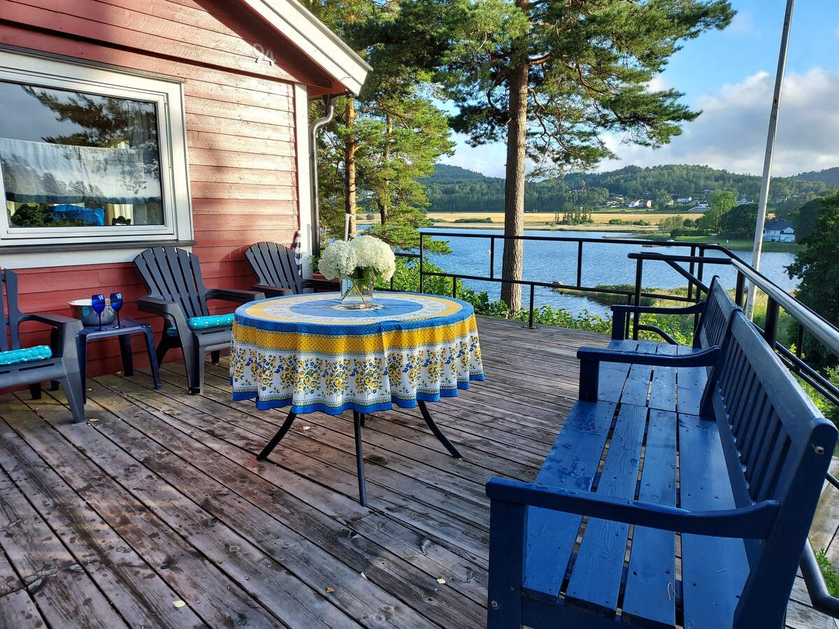 A wooden deck features a round table adorned with a colorful tablecloth and fresh flowers. Two blue chairs and a bench are positioned for seating. The backdrop includes scenic views of the water and surrounding greenery, contributing to a peaceful outdoor space.