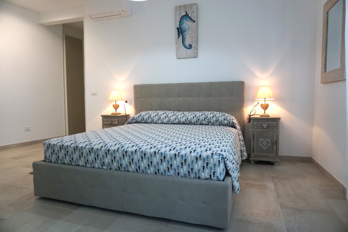 A spacious bedroom features a comfortable bed with a patterned coverlet. Bedside tables with lamps provide soft lighting, while a decorative seahorse artwork is displayed above the headboard. The room is finished with neutral tones and modern flooring, creating a calming atmosphere.