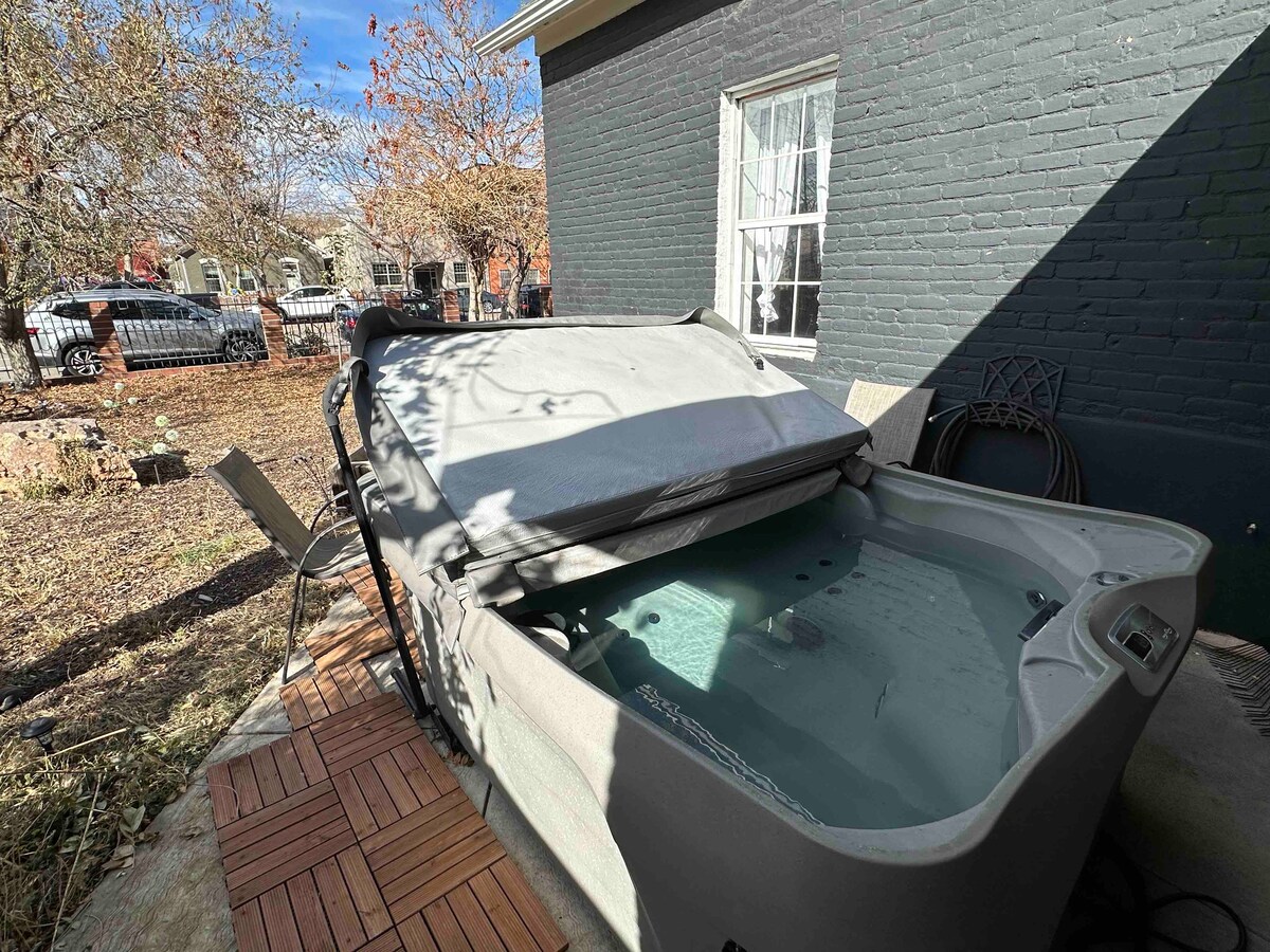 A hot tub sits in a private outdoor area, surrounded by natural scenery. A cover rests partially over the tub, allowing water to be visible inside. The space is designed for relaxation, with a nearby seating area and sunlight filtering through surrounding trees.