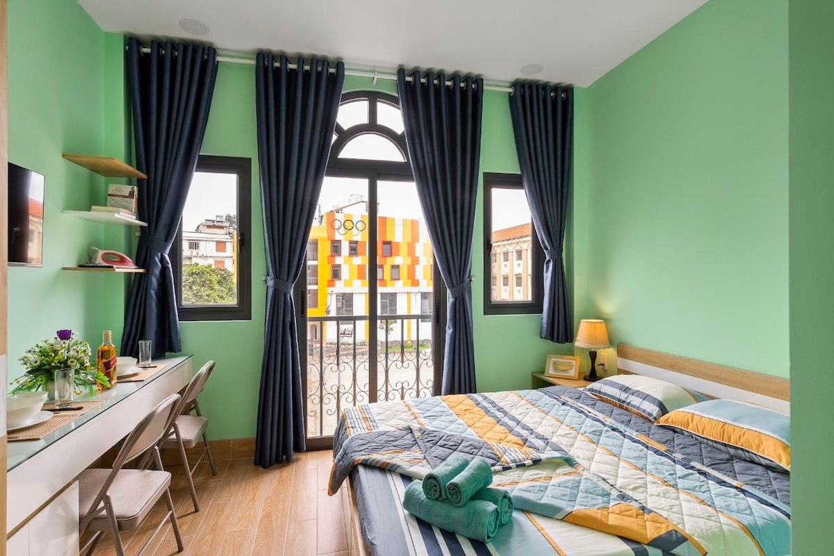 A bright sleeping area features green walls and large windows with patterned curtains. A spacious bed with patterned bedding is complemented by rolled towels and a bedside lamp. A workspace with a table and chairs is positioned near the window, offering a view of the vibrant exterior.