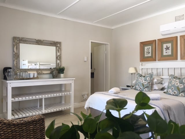 A bright guest room features a spacious bed adorned with decorative pillows and soft linens. A large mirror with an ornate frame reflects the serene decor. A simple wooden console table holds glasses and a kettle, while greenery adds a natural touch.