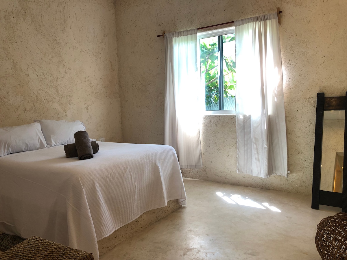 A spacious bedroom features a large bed with white bedding and accent towels. Natural light enters through a nearby window, which is covered by sheer white curtains. A simple wooden mirror stands against the wall, reflecting the light and inviting atmosphere.