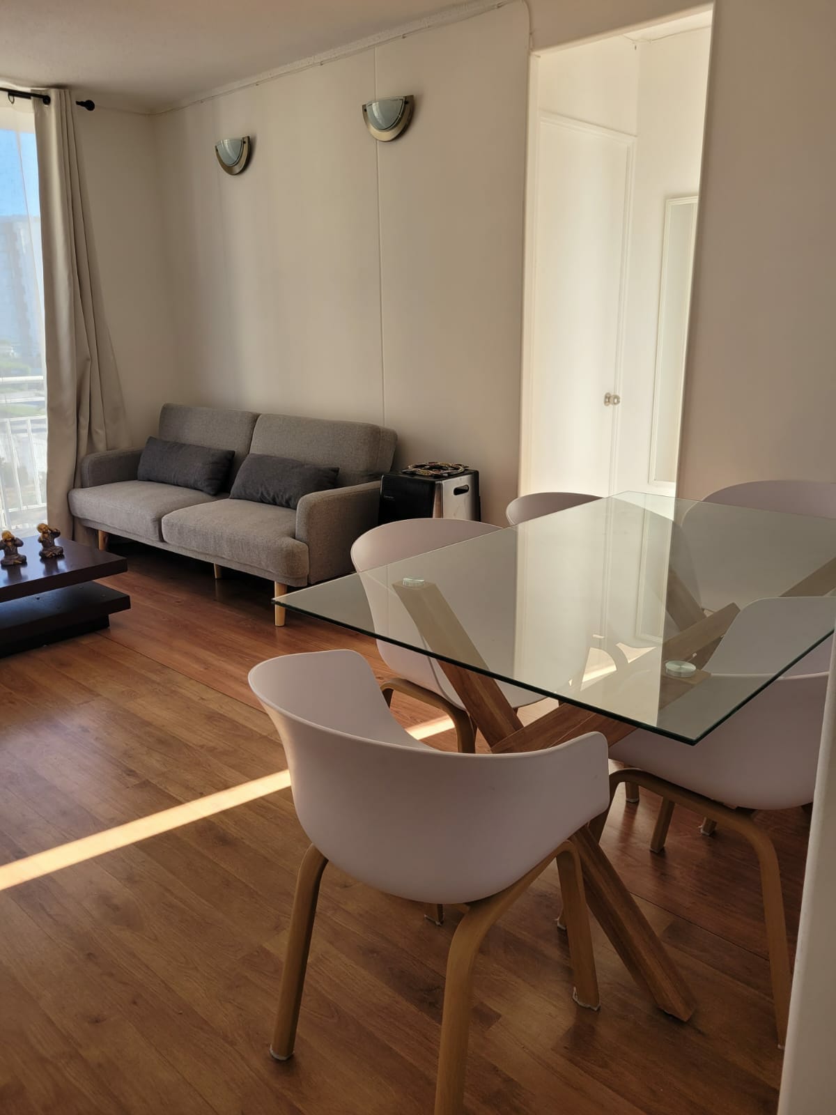 A bright dining area features a glass table surrounded by four light-colored chairs. Natural light filters through a nearby window, illuminating the space. A gray sofa offers additional seating, complemented by a small coffee table. Light hardwood flooring enhances the room's openness.