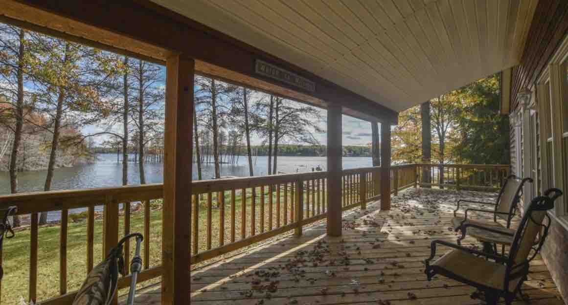 The outdoor deck features a wooden railing overlooking the tranquil waters of Big Bear Lake. Several chairs are positioned for relaxation, with scattered leaves on the wooden floor, surrounded by trees that enhance the scenic lakefront view.