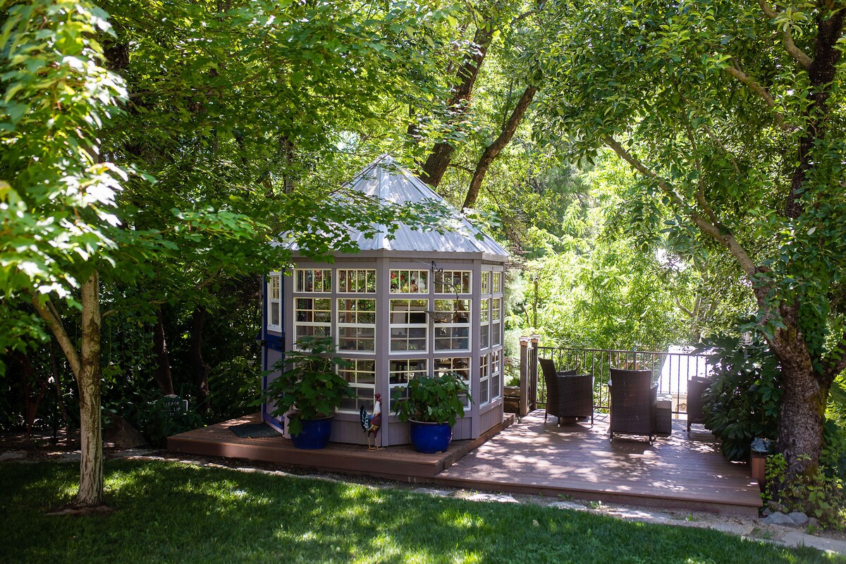 A charming gazebo is framed by lush greenery and surrounded by potted plants. A well-maintained deck features cozy seating, offering a peaceful space to relax. The setting showcases a tranquil garden atmosphere, with soft sunlight filtering through the trees.