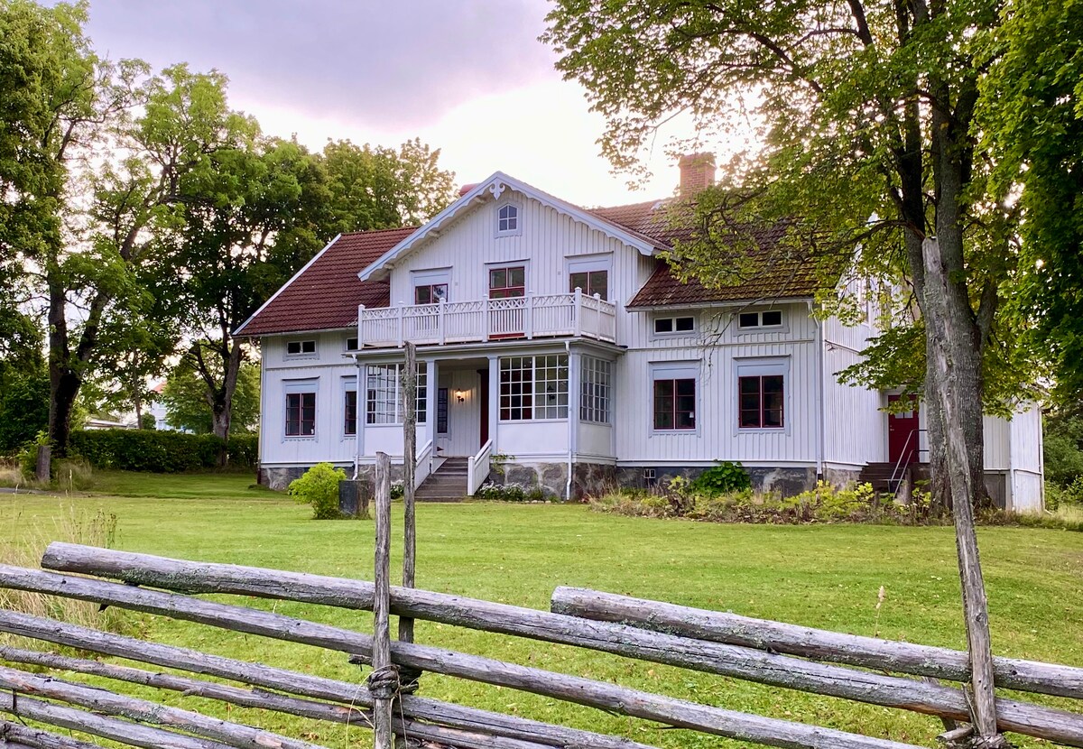 A traditional two-story white house is situated on a grassy landscape, framed by lush trees. A wide staircase leads to the entrance, which is adorned with a covered balcony. A rustic wooden fence runs parallel to the property, enhancing the tranquil countryside setting.