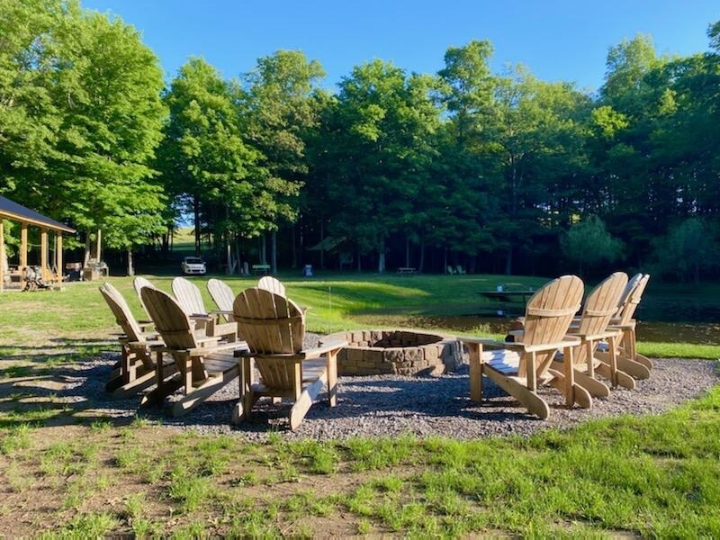 A circular arrangement of wooden Adirondack chairs encircles a brick fire pit, situated on a grassy area next to a koi pond. Tall trees provide shade in the background, contributing to a serene woodland atmosphere.