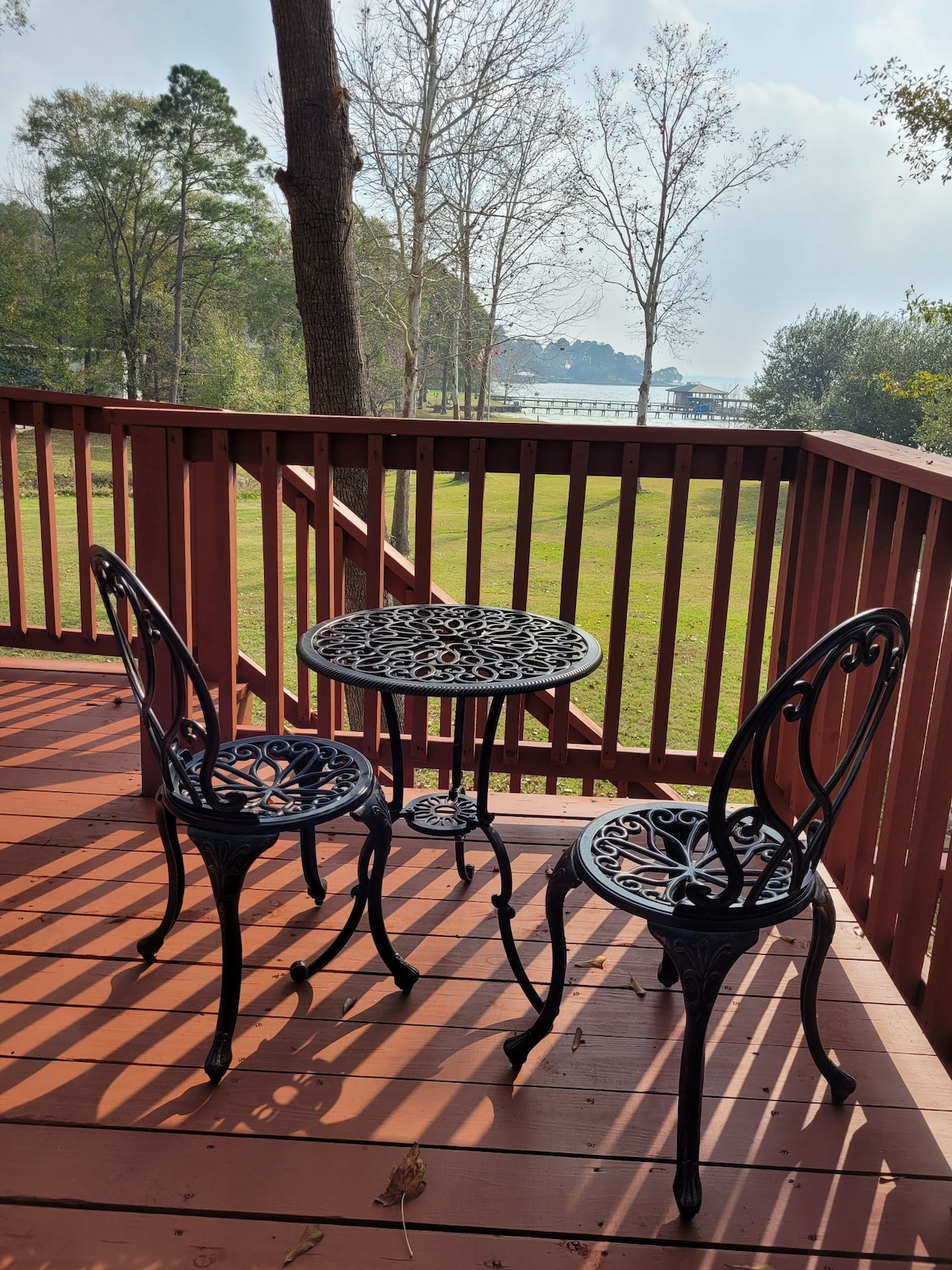 A small outdoor seating area features two decorative metal chairs and a matching round table, positioned on a wooden deck. The serene view includes a glimpse of the lake through the trees, with a peaceful green lawn complementing the scene.