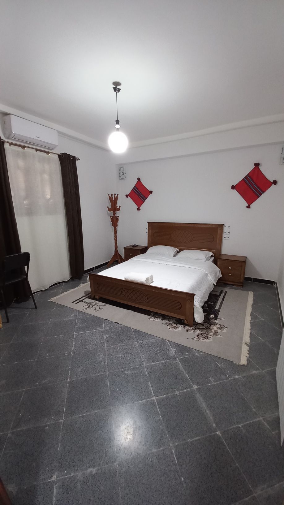 A spacious sleeping area features a wooden bed with a light blanket and decorative towels. Two side tables are positioned next to the bed. Wall art reflects local culture, and a chair and small table are included. Natural light enters through a partially draped window.