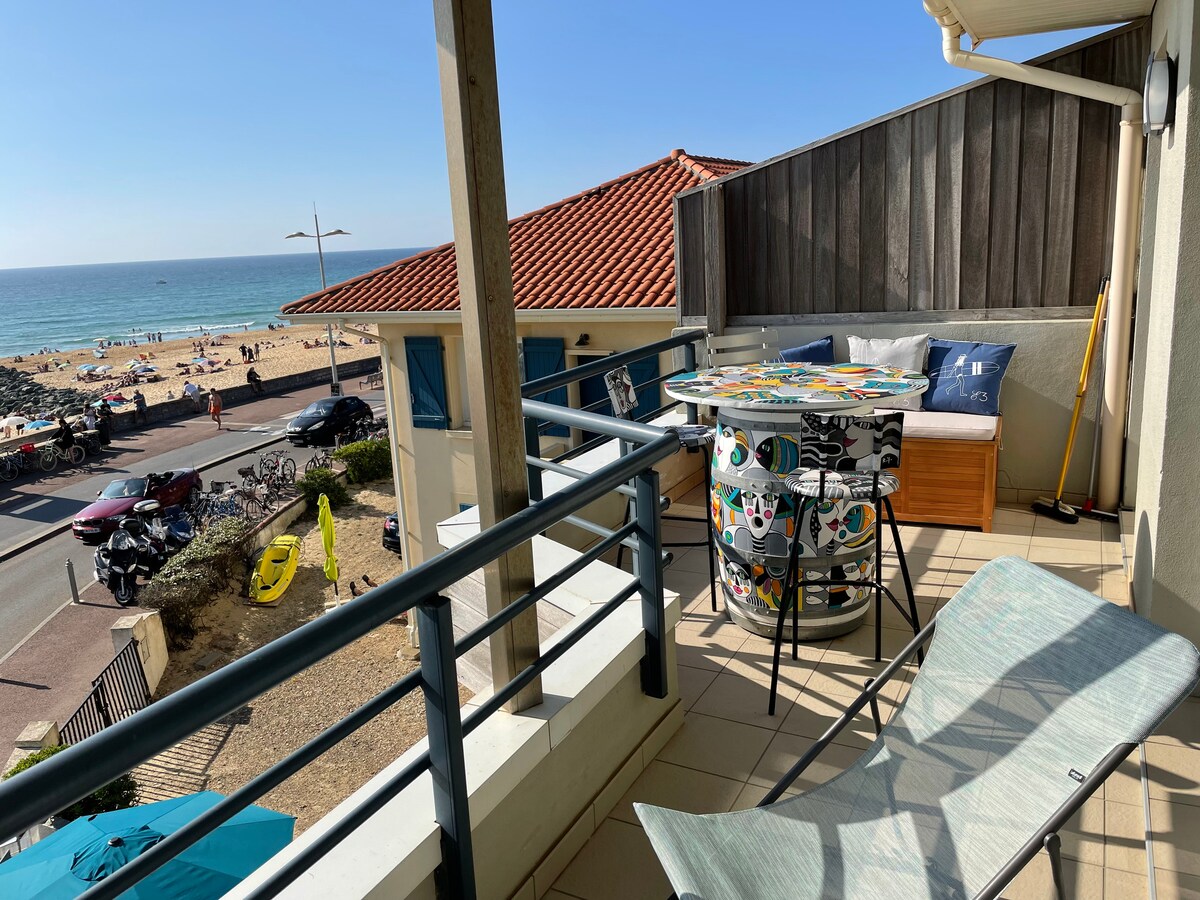 A balcony is presented with comfortable seating and a colorful table. Ocean views are observable, with the beach and surfers in the distance. Sun loungers are arranged for relaxation, while vibrant beach activities unfold nearby.