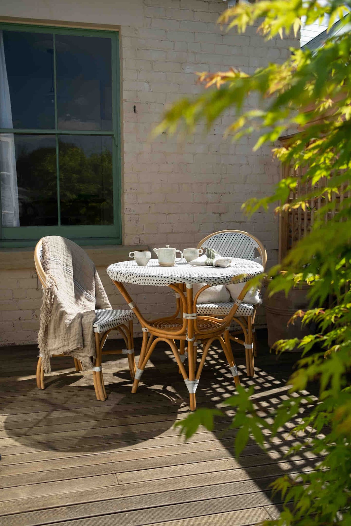 A round table is set with a tea service, surrounded by three woven chairs. A cozy throw drapes over one chair. The area is illuminated by sunlight filtering through a nearby window, with green foliage framing the scene.