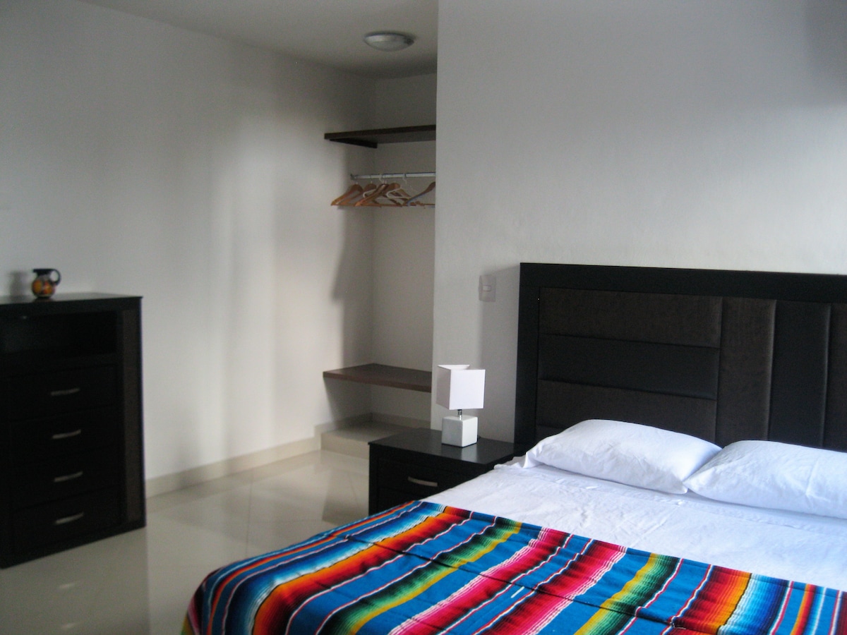 A spacious bedroom is featured, showcasing a large bed adorned with a colorful blanket. An adjacent nightstand holds a lamp, while a closet with hangers is visible. The room is softly illuminated by natural light reflecting off the glossy tile floor.