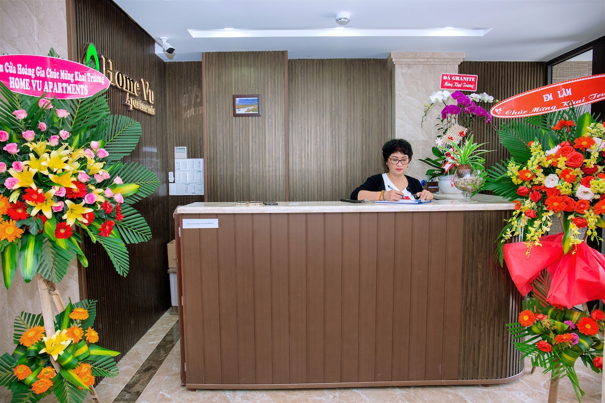 A reception area is present with a wooden front desk, decorated with colorful floral arrangements on either side. The backdrop features light brown paneling, and a friendly staff member is positioned behind the desk, welcoming guests with a warm smile.