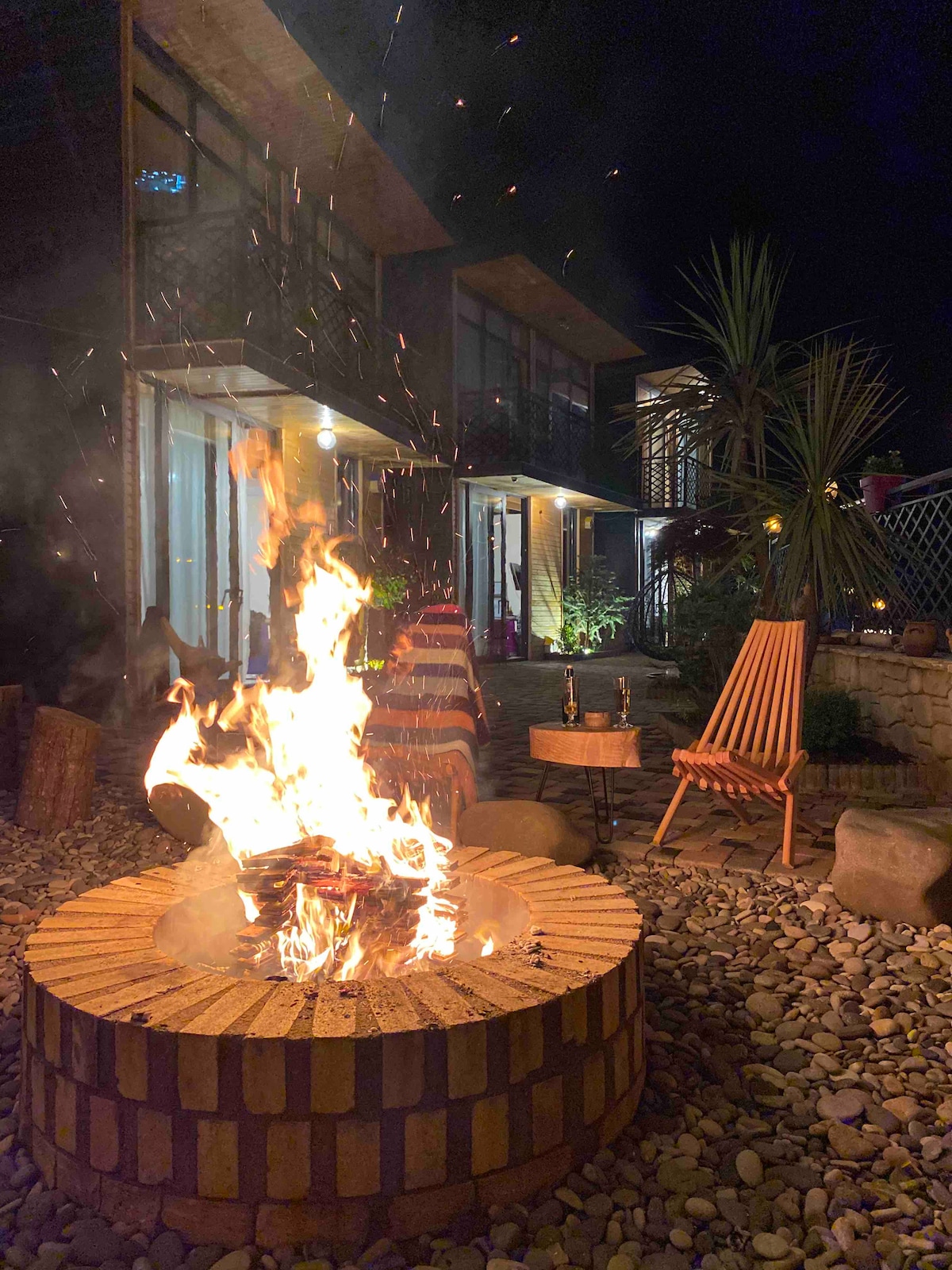 A fire pit is surrounded by a circular stone border, with flames dancing and sparks rising into the night air. Nearby, a wooden chair offers a comfortable seating option. Soft lighting illuminates the exterior of the building, highlighting plants and the inviting ambiance.