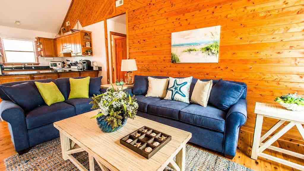 An inviting living area features a navy blue sofa adorned with cream and green pillows. A light-colored coffee table rests in front, displaying a floral arrangement and a decorative tray. Natural wood walls contribute to a cozy ambiance, with a glimpse of the kitchen visible in the background.