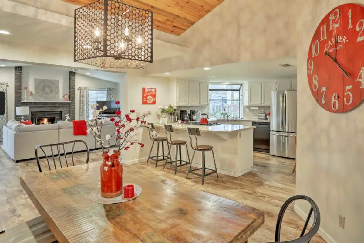 An open concept dining area features a rustic wooden table with seating for six. A contemporary chandelier hangs above, while a decorative vase with flowers adds a pop of color. In the background, a modern kitchen is visible, showcasing sleek cabinets and stainless steel appliances.