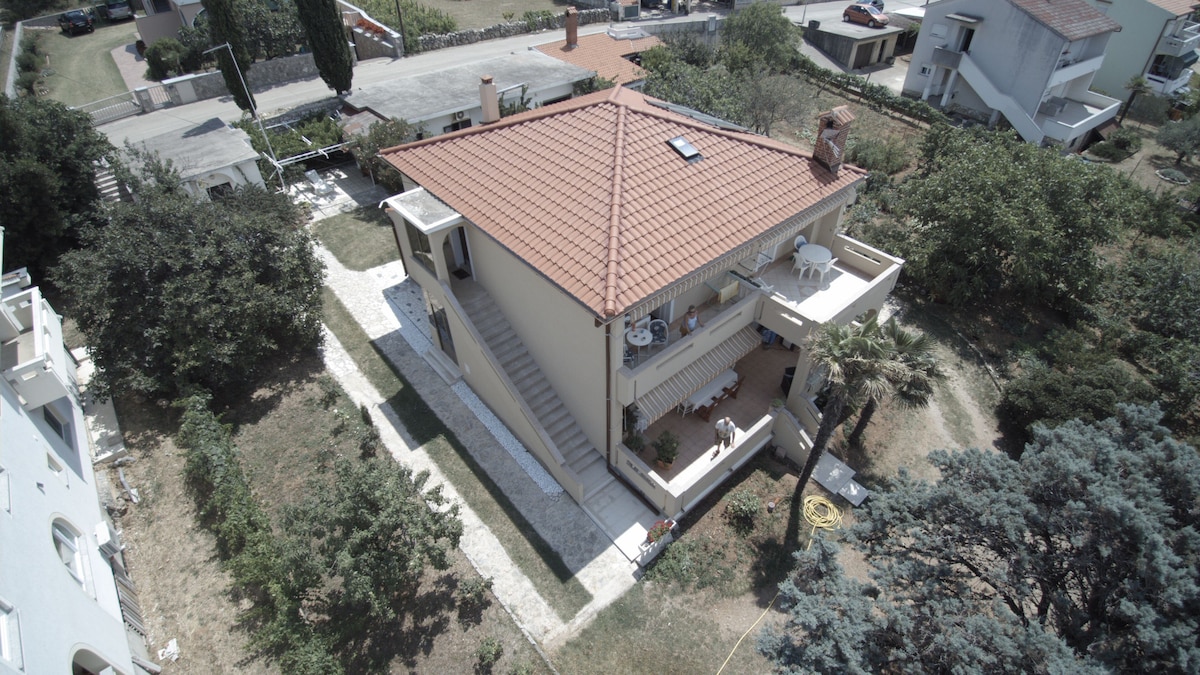 An aerial view showcases the family property with a charming terrace. The apartment is situated above the owner's house, surrounded by greenery. A pathway leads up to the entrance. Nearby vehicles are visible parked along the street.