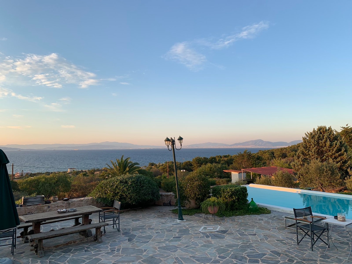 A panoramic view showcases the sea and distant mountains, framed by lush greenery. A stone patio features a wooden dining table surrounded by black chairs, while a swimming pool reflects the sky. A lamp post adds charm to the serene outdoor setting.