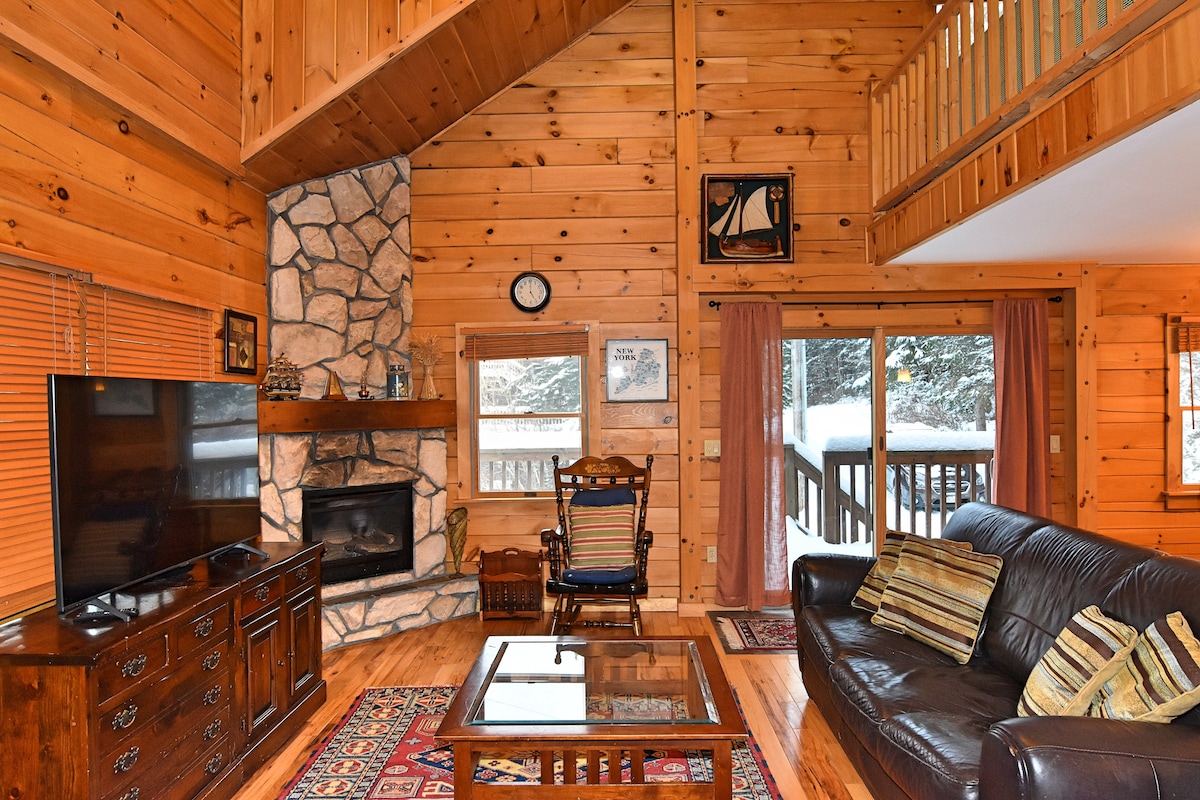 A cozy living area features a stone fireplace, dark leather seating, and a wooden coffee table. Large windows provide views of the outdoors, with natural light filling the space. A vintage rocking chair and decorative artwork add character to the warm wooden interior.