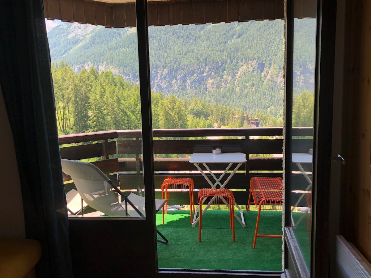 A balcony scene is visible through sliding glass doors, featuring a small table and four colorful chairs on green turf. A sun lounger is present, surrounded by a scenic view of mountains and trees, creating a serene outdoor space.