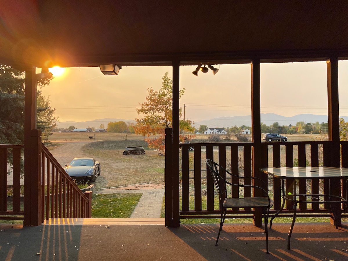 A covered outdoor porch is seen at sunset, revealing a peaceful view of the landscape. A small dining table is positioned next to iron chairs, while a vehicle is visible in the driveway. The warm glow of the sun casts a serene atmosphere over the area.