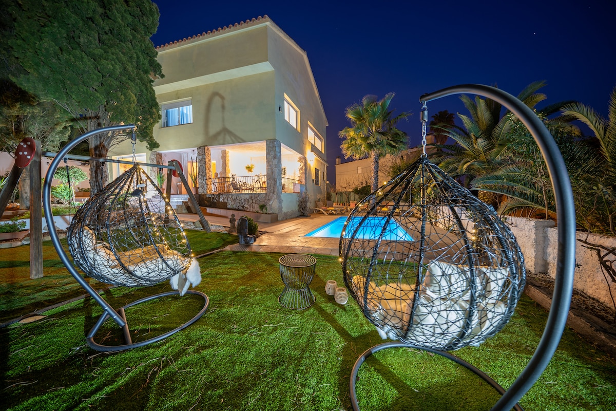 Two hanging chairs are placed on a grassy area, facing a private pool illuminated under the night sky. The villa's façade can be seen in the background, showcasing its modern architecture surrounded by palm trees and lush greenery.