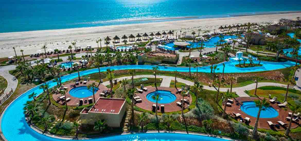 A panoramic view captures multiple swimming pools winding through the resort, surrounded by lush greenery and sun loungers. The ocean sparkles in the background, with palm trees lining the shore, providing a serene coastal atmosphere and inviting relaxation.