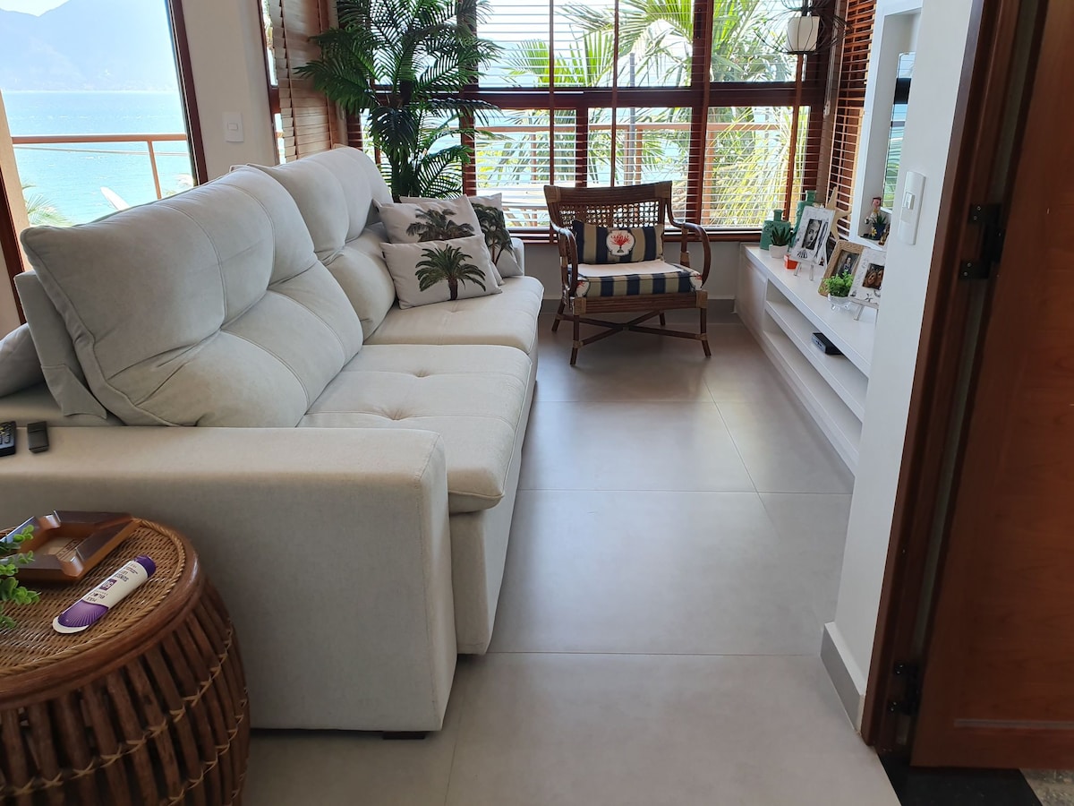 A bright living area features a light-colored sofa paired with a wooden accent chair. Natural light filters in through large windows, highlighting the greenery outside. A modern TV unit is positioned against the wall, adorned with a few personal items and framed photos.