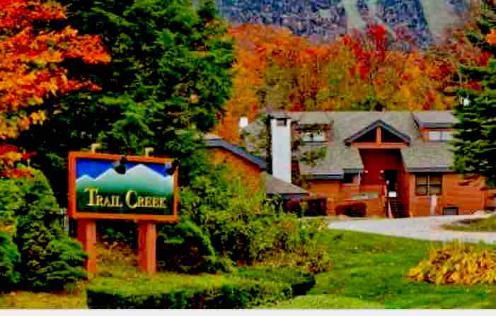 The exterior of the Trail Creek complex is showcased, surrounded by vibrant autumn foliage. A welcoming sign reading 'Trail Creek' is prominently displayed near a manicured lawn. The building features a combination of rustic wooden elements and contemporary architecture, set against a mountainous backdrop.