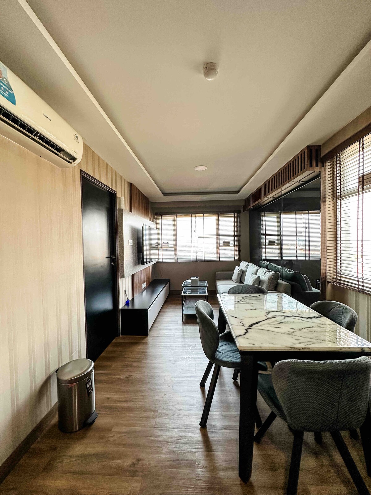 A sleek dining area features a marble-topped table surrounded by upholstered chairs. A large window with vertical blinds allows natural light to fill the space, while a flat-screen TV is mounted on a wooden console across from the table, creating a cozy atmosphere.