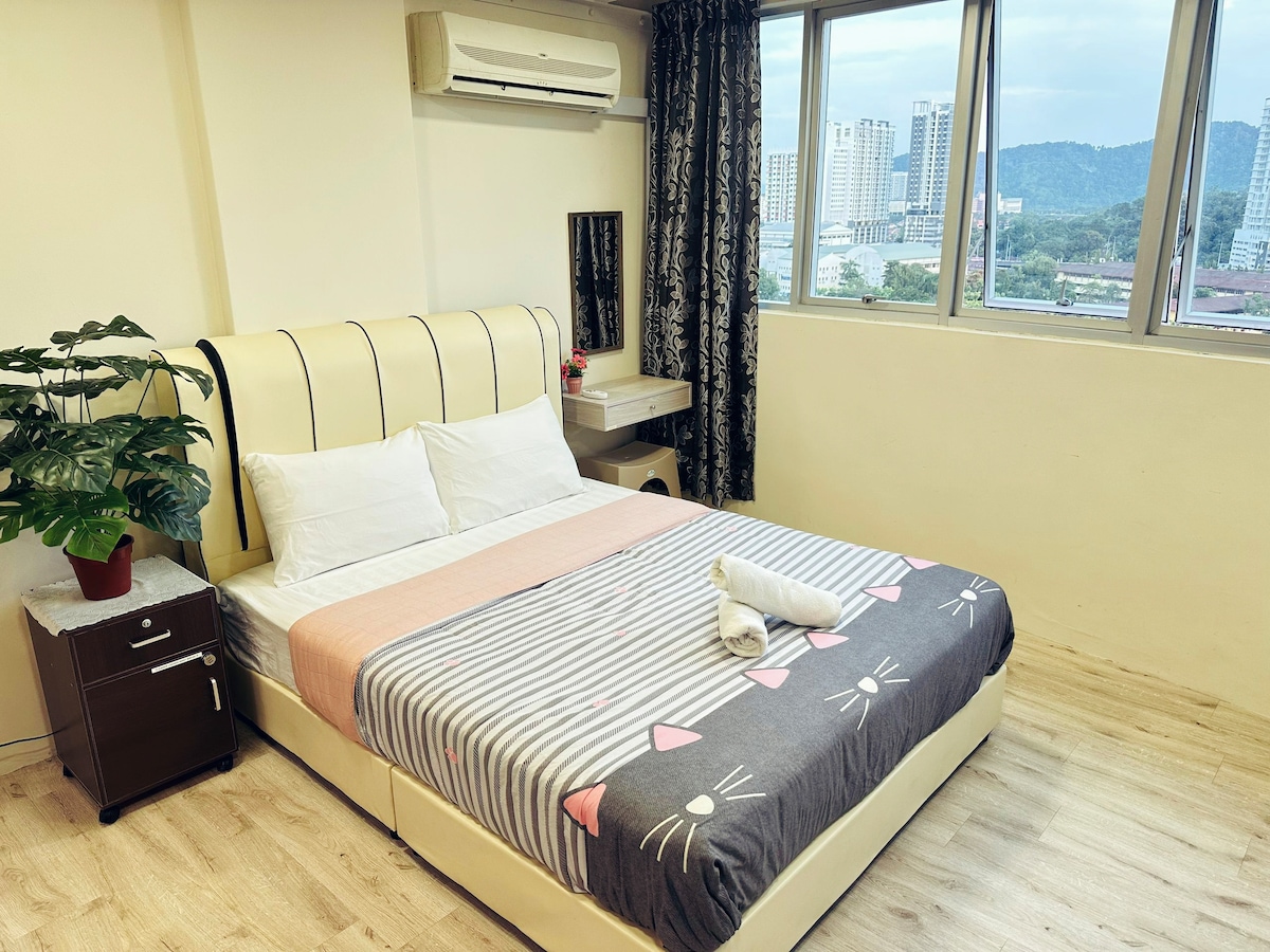 A well-furnished sleeping area features a spacious bed with decorative linens and a soft grey blanket. Natural light enters through large windows, providing a view of the surroundings. A small bedside table and a potted plant add subtle touches to the inviting space.