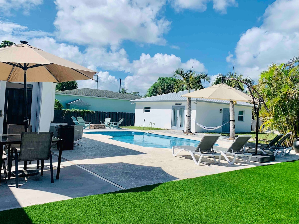A sunny outdoor area features a private heated pool surrounded by lounge chairs and an umbrella-shaded dining table. Lush green grass enhances the landscape, while palm trees provide a tropical ambiance. Two houses are visible in the background, contributing to the inviting setting.