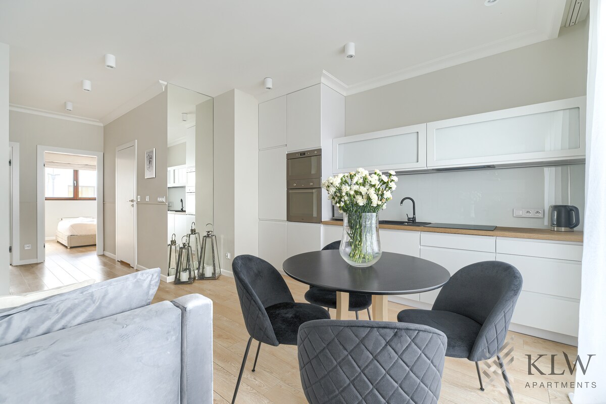 A modern kitchen is visible with sleek white cabinetry and stainless steel appliances. A round dining table is accompanied by four black chairs. Fresh flowers are arranged in a clear vase on the table. The open layout allows for natural light to fill the space.
