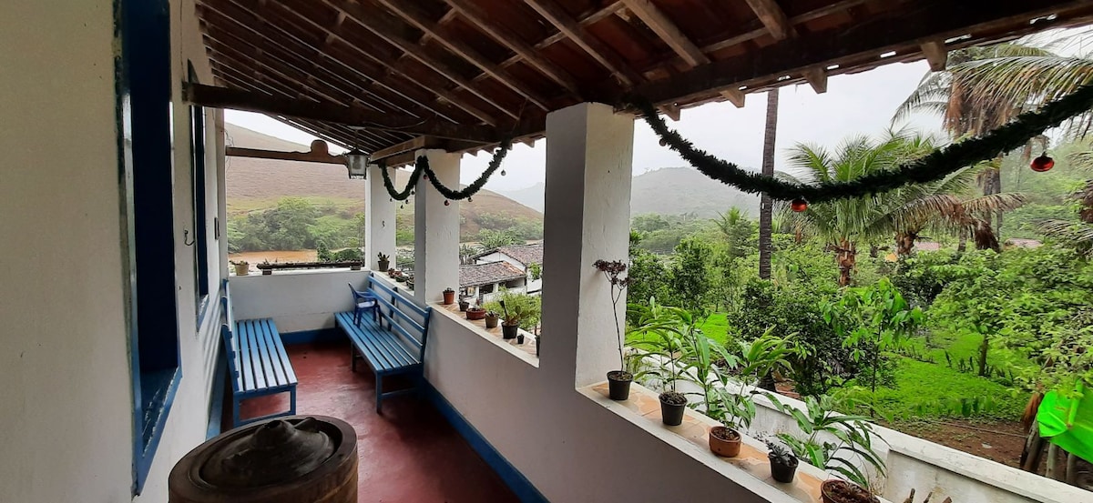 A covered porch area features wooden benches along the walls, adorned with potted plants. Scenic views of lush greenery and distant hills are visible beyond the railing. The space is complemented by a wooden barrel, enhancing the rustic charm.