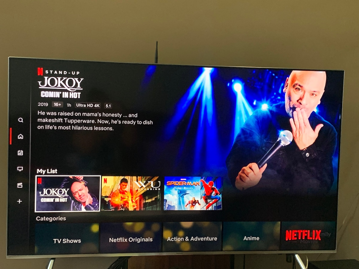 A large television screen displays the Netflix interface, featuring a stand-up comedy special titled 'Jokoy: Comin' In Hot'. The screen shows vibrant visuals with colorful stage lighting and a performer holding a microphone, surrounded by icons for categories and options on the user interface.