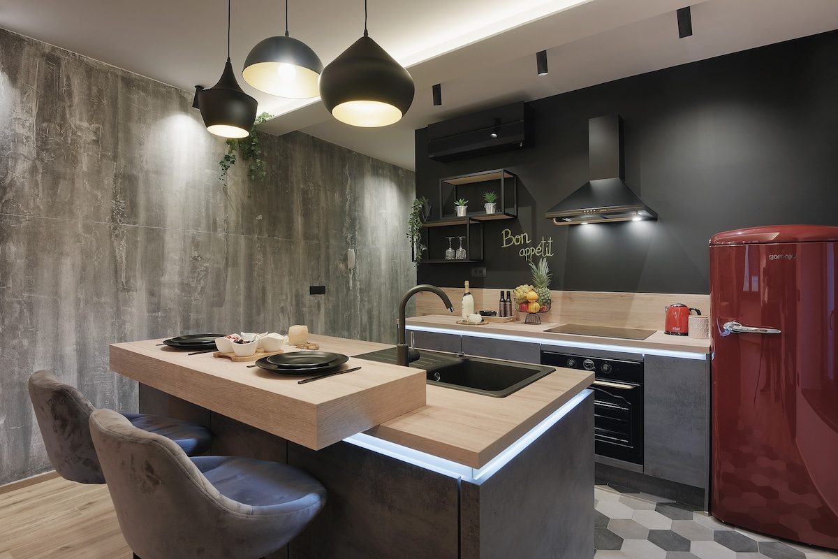A modern kitchen features a wooden island with seating for two, set against stylish concrete walls. Black cabinetry complements the design, while a vibrant red refrigerator adds a pop of color. Soft lighting enhances the overall ambiance and usability of the space.