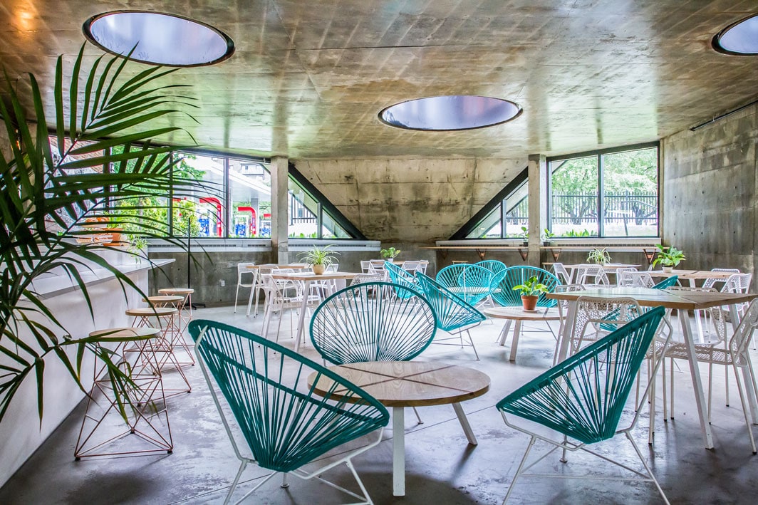 A spacious communal area features modern seating arrangements, including teal-colored chairs and light wooden tables. Large circular windows allow natural light to fill the space, while potted plants add a touch of greenery. The concrete walls provide a minimalist backdrop, enhancing the contemporary aesthetic.