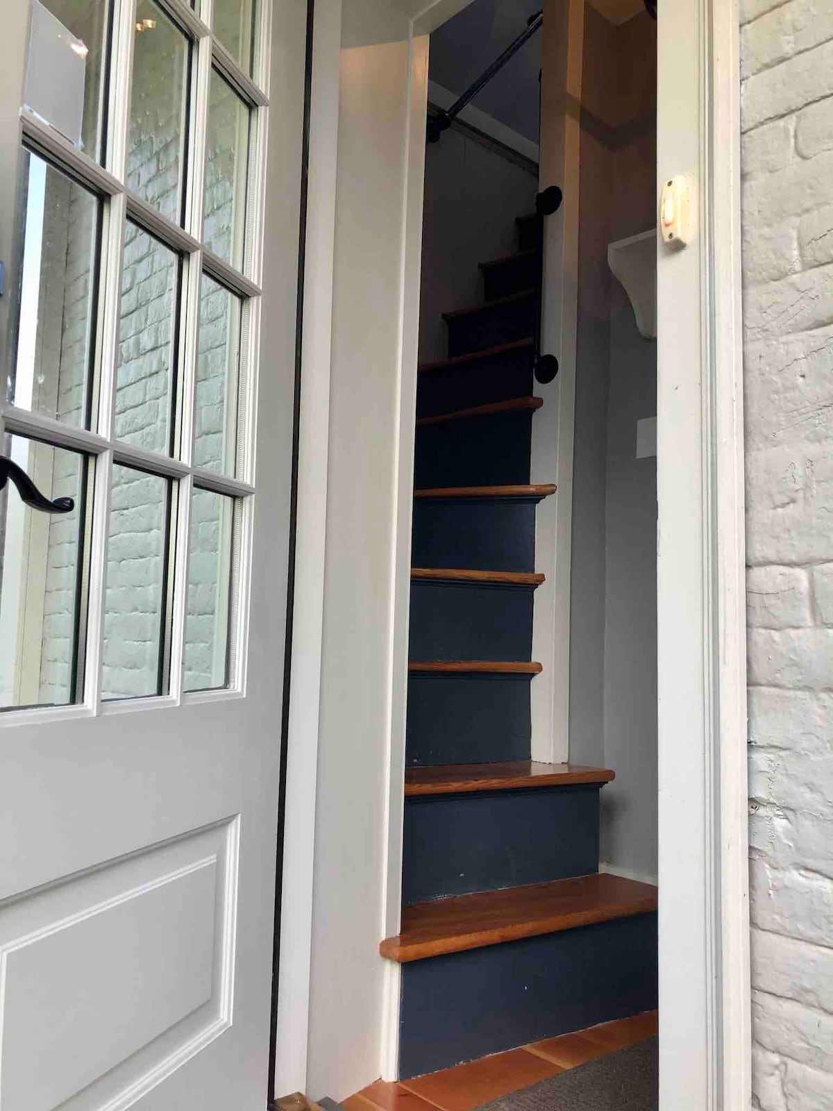 A steep staircase is seen leading up from a welcoming entrance. The staircase features wooden steps and a handrail, flanked by light-colored walls, with natural light illuminating the space from the entrance.