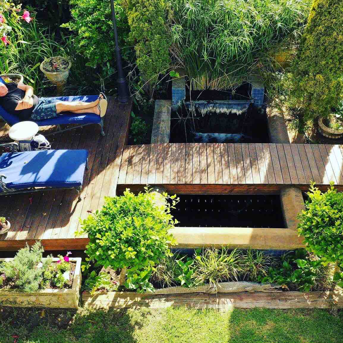 A serene outdoor space features a wooden deck surrounded by lush greenery and potted plants. A tranquil water feature is visible, enhancing the area’s peaceful atmosphere. A lounge chair offers a spot for relaxation under soft natural light, integrating nature with comfort.