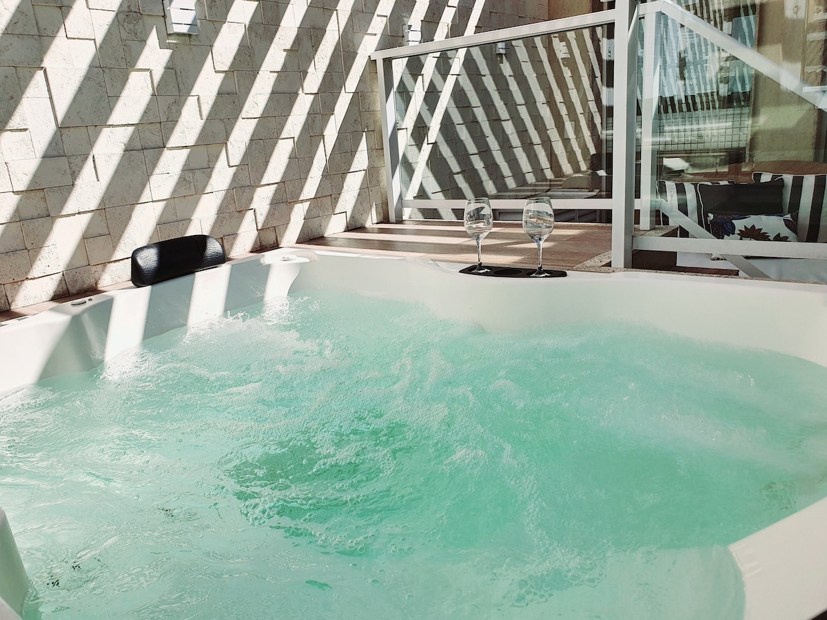 An outdoor jacuzzi is visible, surrounded by wooden decking and glass railing. The water appears inviting, with gentle bubbles creating ripples. Two wine glasses are positioned on the edge of the jacuzzi, suggesting a relaxed atmosphere, enhanced by diagonal light patterns from the overhead structure.
