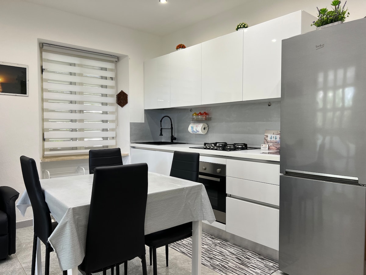 A modern kitchen featuring white cabinets and a sleek countertop is shown. A dining table with black chairs is set up in the center. Essential appliances, including a silver refrigerator and a gas stove, are visible, with natural light coming through the window equipped with roller blinds.