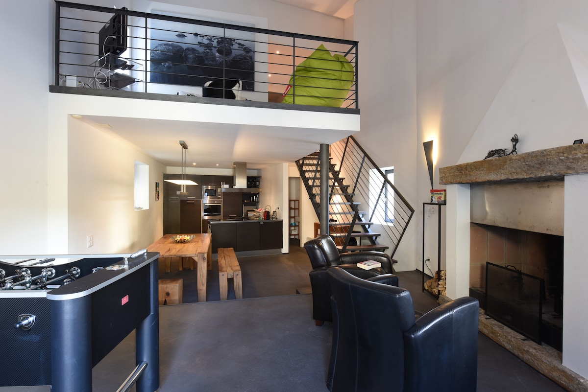 An open-plan living space features a modern kitchen with sleek cabinetry and a wooden dining table. Two black armchairs are positioned near a stone fireplace. A staircase leads to a loft space, while vibrant decorative elements add interest to the neutral walls.